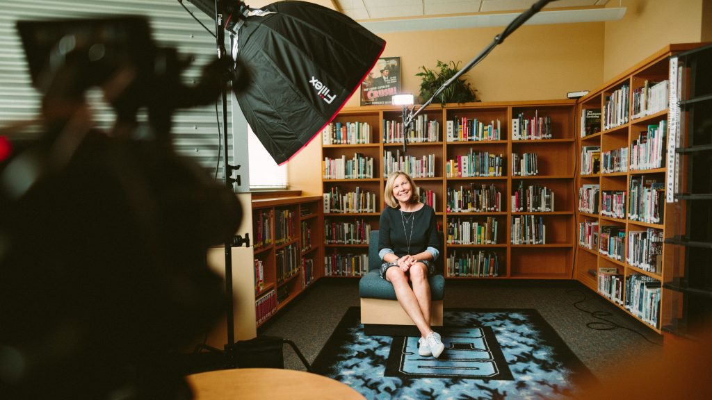 woman sitting in front of a camera with a bookshelf in the background