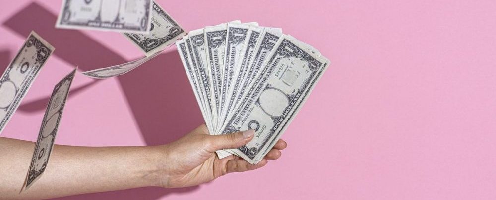 hand holding dollar bills in front of a pink wall