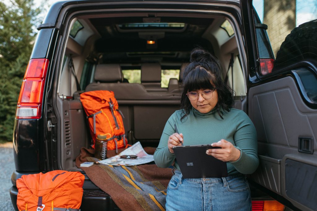 woman in leaning against back of open car hatch looking at tablet