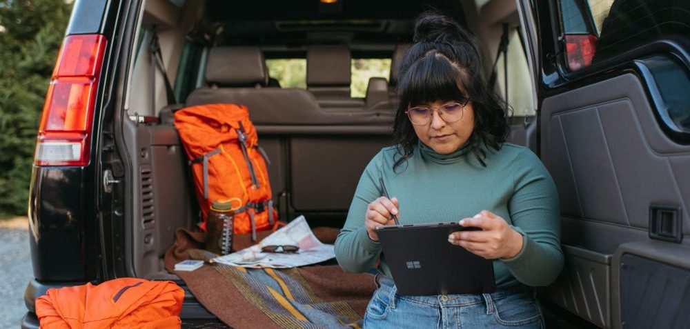 woman in leaning against back of open car hatch looking at tablet