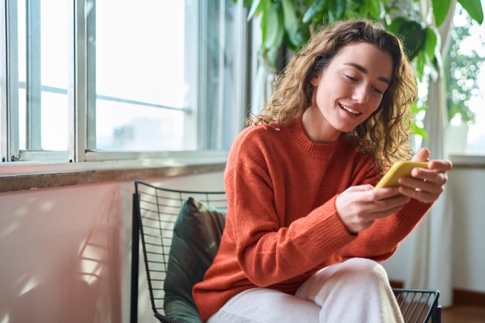 Smiling young woman sitting on chair holding mobile phone using cell at home.