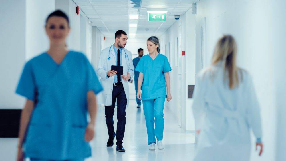 Surgeon and Female Doctor Walk Through Hospital Hallway, They Consult Digital Tablet Computer while Talking about Patient's Health. Modern Bright Hospital with Professional Staff.