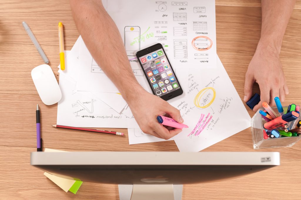 A hand making notes on a piece of paper on a desk with a computer monitor and a phone